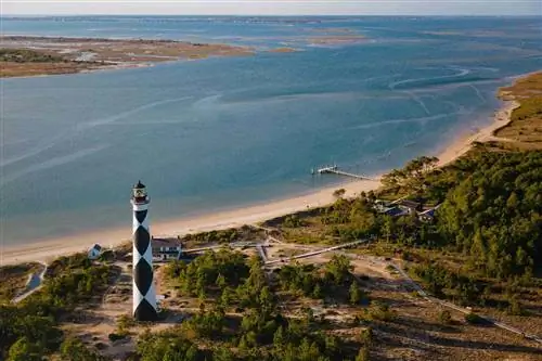 Вид с воздуха на национальное побережье Кейп-Лукаут, маяк Кейп-Лукаут и музей Кварталов хранителей, офисы Службы национальных парков, сувенирный магазин и пристань с заливом Барден, отмелями Шеклфорд и островом Харкерс вдалеке и зеленой листвой вдоль береговой линии.
