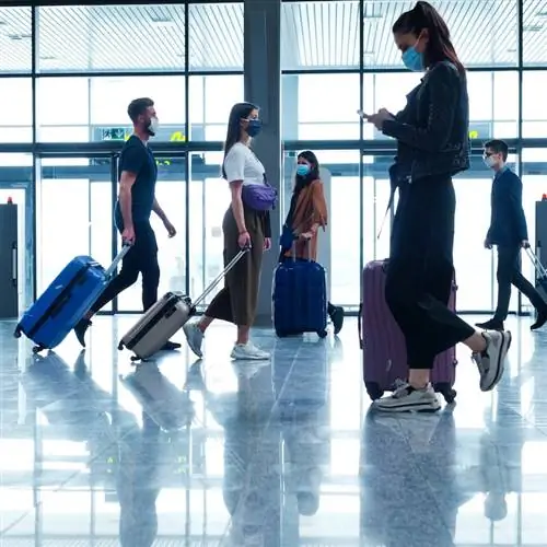 Люди, путешествующие на самолете во время COVID 19, в масках N95, несут багаж в терминале аэропорта