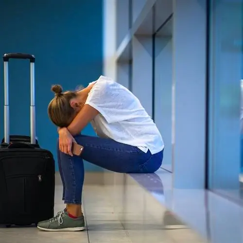 Путешественница сидит на земле в аэропорту перед своим чемоданом на колесиках, положив голову на колени