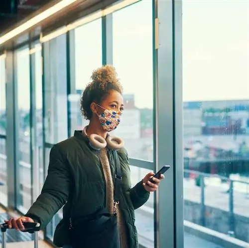 Путешественница держит чемодан у ворот аэропорта, в маске