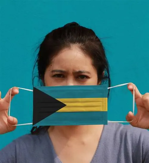 Женщина с флагом Багамских островов на гигиенической маске в руке и подняла лицо на голубом фоне. Крошечные частицы или вирусная корона или защита от Covid 19. Концепция борьбы с болезнью. (Женщина с флагом Багамских островов на гигиенической маске в ее