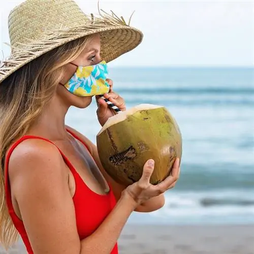 Женщина-путешественница пьет кокосовую воду из кокоса с маской для лица