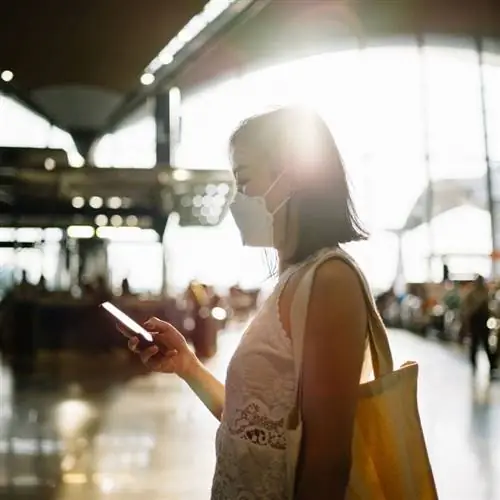 Молодая азиатка в маске, проверяя телефон в аэропорту