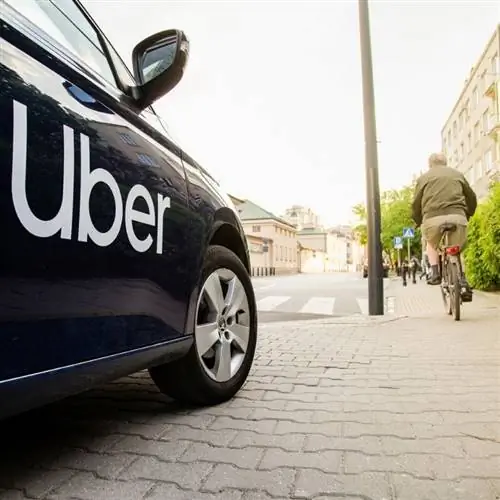 Сторона черного автомобиля Uber, припаркованного на тротуаре, когда велосипедист проезжает мимо в неустановленном месте Сторона черного автомобиля Uber, припаркованного на тротуаре, когда велосипедист проезжает мимо в неустановленном месте
