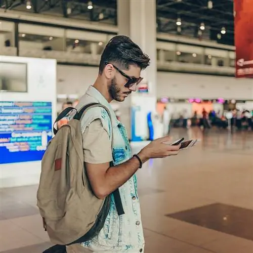 Молодой путешественник-мужчина проверяет свой телефон в аэропорту, держа в руках паспорт и посадочный талон