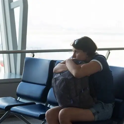 Женщина выглядит уставшей в аэропорту, прислонившись к собственному рюкзаку перед международным путешествием