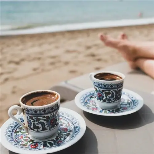 Роскошный пляжный отдых для двоих в Турции. Две чашки турецкого кофе, женские ноги, песок и Средиземное море на заднем плане. Романтический отдых для двоих. Летний день или утро на побережье Анталии