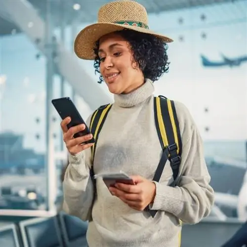 Женщина-путешественница проверяет свой телефон, улыбаясь в аэропорту