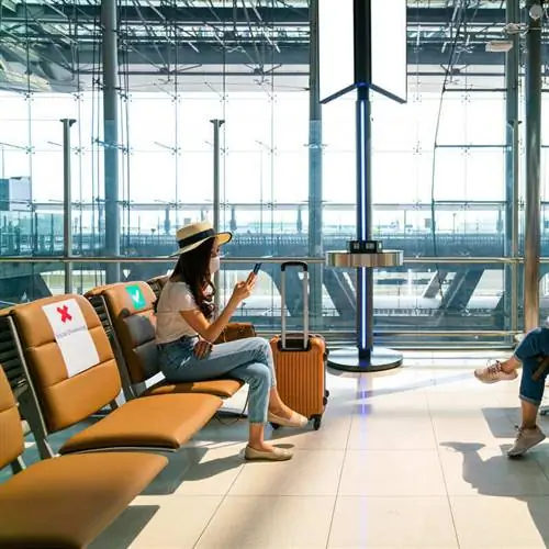 Молодая путешественница смотрит на свой телефон, надевая маску в аэропорту перед авиаперелетом