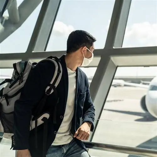 Авиапассажир в маске наблюдает за самолетом через стеклянный фасад терминала аэропорта, концепция международного путешествия