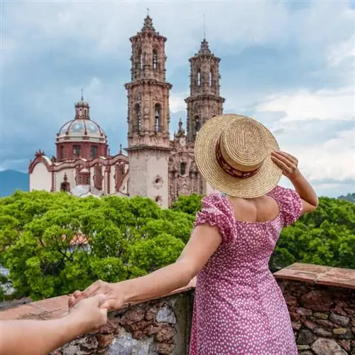 Молодая туристка в розовом платье и соломенной шляпе позирует фотографу, держа за руку своего партнера, смотровую площадку в Таско, Пуэбло Магико в Мексике