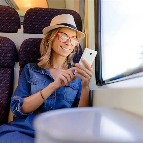 Молодая путешественница проверяет свой телефон, улыбаясь во время поездки на поезде, путешествие на поезде