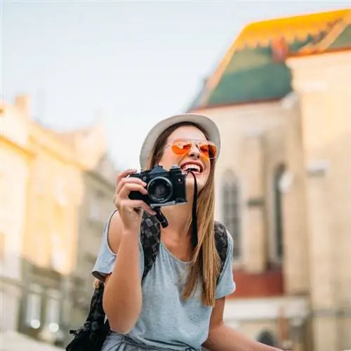 Молодая улыбающаяся женщина-фотограф держит камеру в руках и фотографирует неустановленный европейский город, смутно видимый на размытом фоне