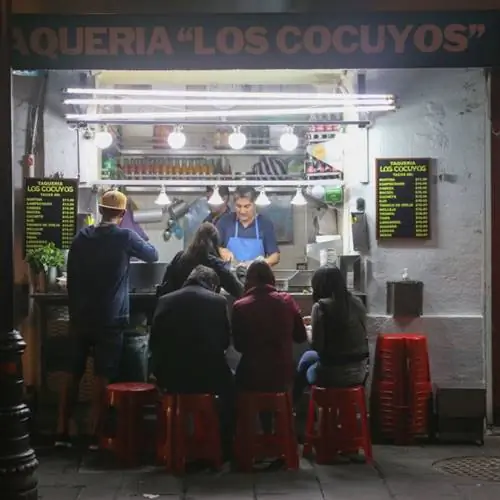 посетители сидят на маленьких красных пластиковых скамейках перед такерией в Мехико и заказывают еду на испанском языке.