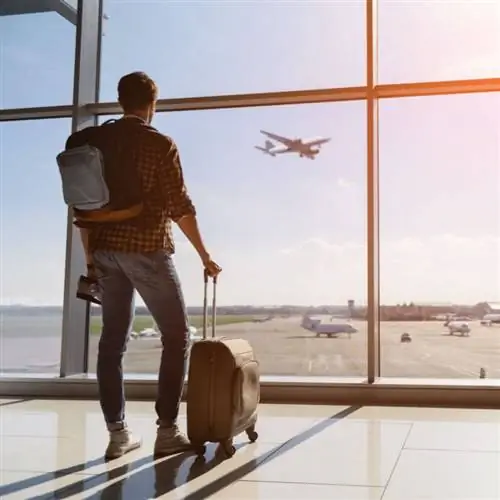 Спокойный турист-мужчина стоит в аэропорту и смотрит на полет самолета через окно. Он держит билеты и чемодан