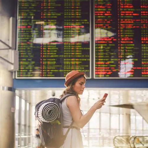 Молодая путешественница проверяет свой мобильный телефон перед информационным табло, на котором изображено несколько отмененных или задержанных рейсов, авиаперелеты