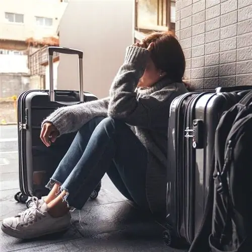 Молодая путешественница выглядит грустной, сидя на полу международного аэропорта рядом со своим багажом