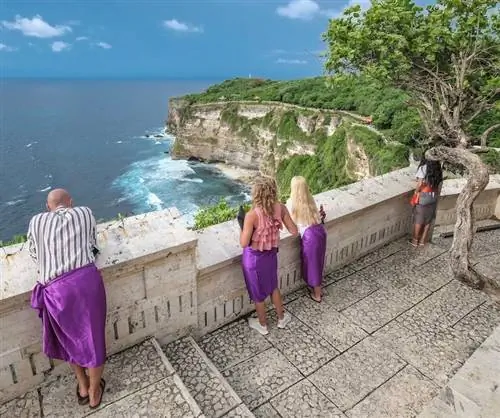 Международные туристы Бали в фиолетовых саронгах в храме с видом на океан в ясный день.