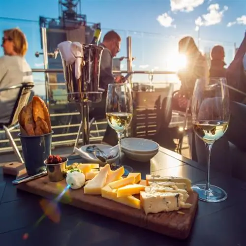 Люди едят сыр и пьют вино в ресторане на крыше во время заката. Стол в ресторане сервирован сырной тарелкой, хлебом и белым вином, полным посетителей.