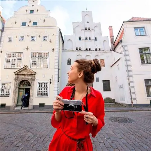 Молодая женщина-фотограф в красной блузке улыбается, любуясь историческими домами Старого города Риги, Латвия, на Балтийском побережье Восточной Европы