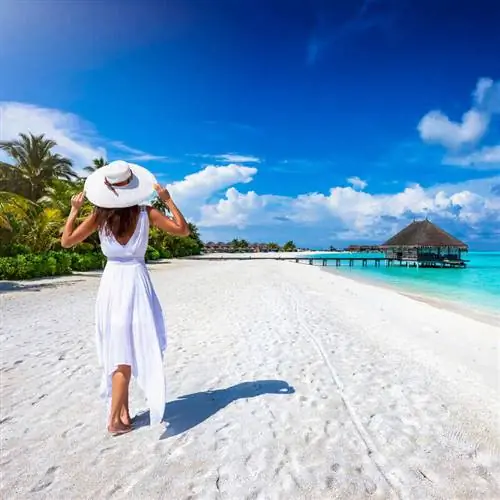 Молодая женщина в белом платье гуляет по тропическому пляжу в неустановленном тропическом месте, вероятно, на Карибах.