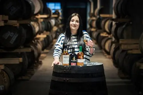 Женщина с длинными черными волосами стоит за тремя стаканами ликера на бочке.
