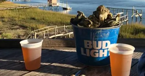 Ведро Bud Light, полное раковин устриц и две чашки пива на деревянном столе на открытом воздухе.