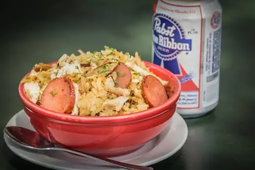 Банка пива рядом с красной миской с рисом, курицей и колбасой