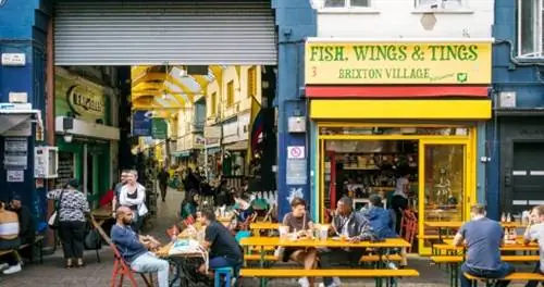 Сцена на улице Брикстон: люди едят за желтыми столами для пикника.