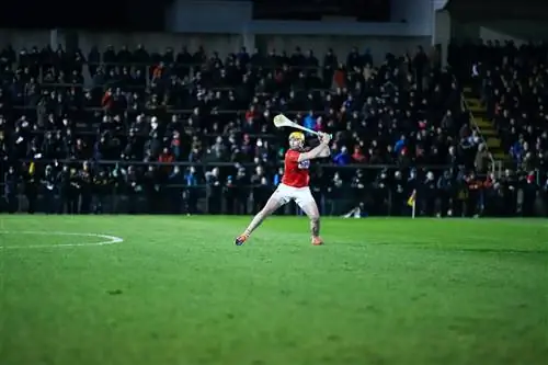 Бросающийся игрок в красном готовится ударить слиотар на глазах у расплывчатой толпы в спортивном комплексе Mallow GAA в Ирландии.