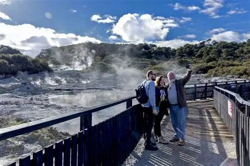 Три человека разного возраста стоят на променаде и позируют для селфи. За ними земля дымится и пузырится