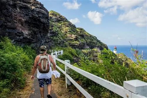 Белый мужчина в бейсболке и шортах идет по тропе маяка Макапуу-Пойнт на Гавайях, а сам маяк находится на дальнем фоне с видом на море.
