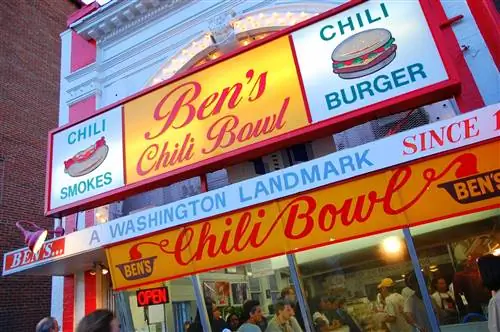 Внешний вид Ben's Chili Bowl, закусочной в округе Колумбия с красочными красными и желтыми винтажными вывесками. За стеклянным окном ресторан переполнен посетителями всех возрастов; выходные в Вашингтоне, округ Колумбия