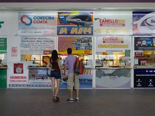 Пара стоит, держась за руки, глядя на кассы на автовокзале.