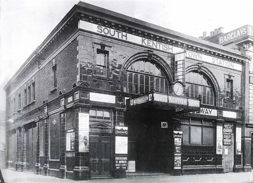 Стареющая черно-белая фотография экстерьера станции метро South Kentish Town; здание кирпичное с черепичным фасадом и арочными окнами над входом.