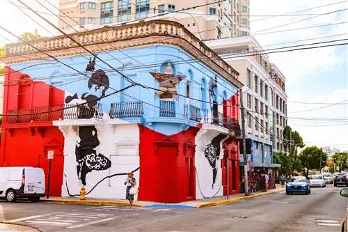 Уличная сцена на улице Calle Loiza в районе Сантурсе в Сан-Хуане, Пуэрто-Рико. Угловое здание украшено флагом Пуэрто-Рико. Вдоль улицы и тротуара можно увидеть машины и людей.