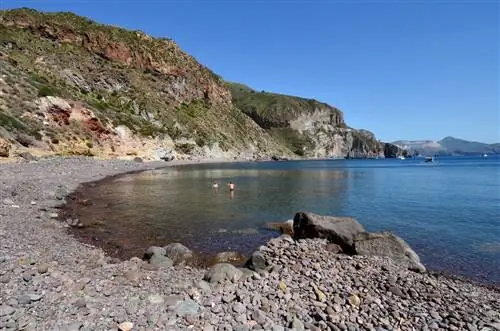 Пляж Валле Муриа: извилистый каменистый пляж, окруженный травянистым мысом. Двое купальщиков плавают на мелководье.