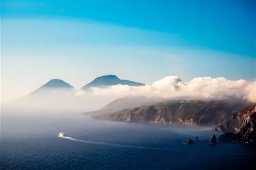 Паром оставляет за собой длинную кривую в море, путешествуя между скалистыми вулканическими островами, над которыми стелется туман.