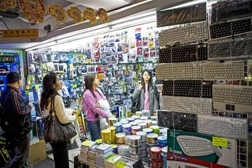 Три человека стоят в хорошо освещенной кабинке со стенами, увешанными электроникой; клавиатуры выставлены на переднем плане. Гонконг.