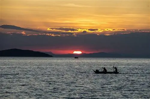 Два рыбака в долбленой каноэ гребут по озеру, когда солнце садится, окрашивая небо в оранжевый и фиолетовый цвета.