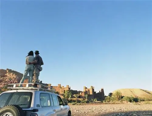 Мужчина и женщина обнимаются на крыше полноприводного автомобиля и смотрят в сторону форта из красного камня на холме.