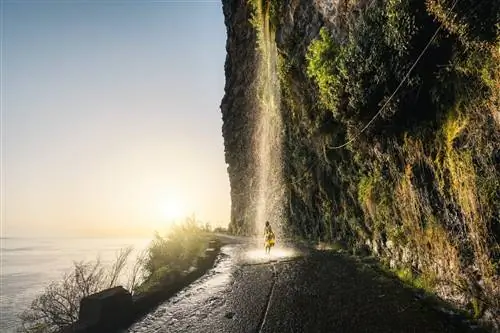 Женщина стоит под водопадом, который льется со скалы посреди дороги на Мадейре, море слева.