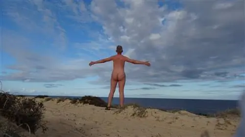 Подняв руки по бокам, обнаженный мужчина стоит на вершине песчаной дюны на нудистском пляже и смотрит на недалекое море; голубое небо над головой имеет красивый водоворот белых и серых облаков