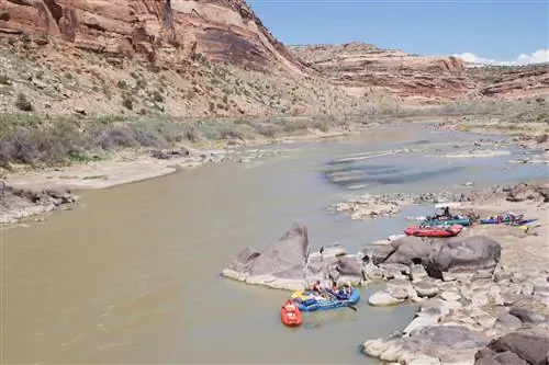 Несколько плотов расставлены по берегу довольно спокойной реки на юго-западе, за которой возвышаются пыльные холмы.