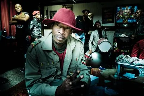 Мужчина в широкополой красной шляпе сидит рядом с камерой в окружении своих товарищей по группе в разных позах.