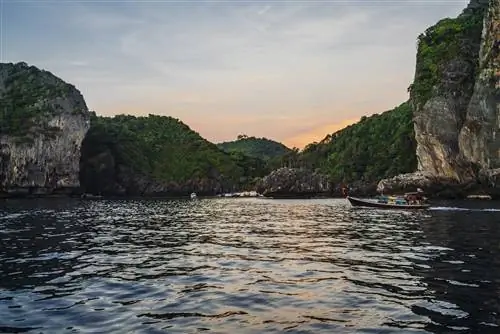Снимок сделан из воды лицом к берегу, усеянному большими скалистыми утесами, покрытыми растительностью. Маленькая красочная рыбацкая лодка плывет мимо