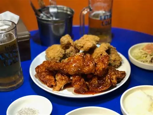 Тарелка корейской жареной курицы, покрытая красным соусом, на синем столе с кружками пива на заднем плане