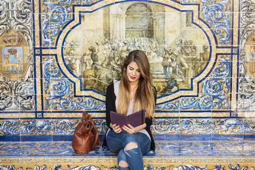 Молодая женщина, читающая вист с цифрового планшета, сидит на площади Испании в Севилье с плиткой на заднем плане.