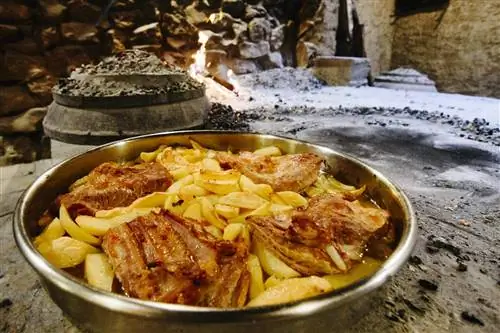 Металлическая посуда с мясом и картофелем, на фоне пепла от камина и крышки из пека.