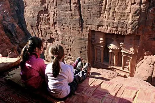 Две молодые девушки сидят на скале и смотрят вниз, в долину, где древний город Петра высечен в красном камне.